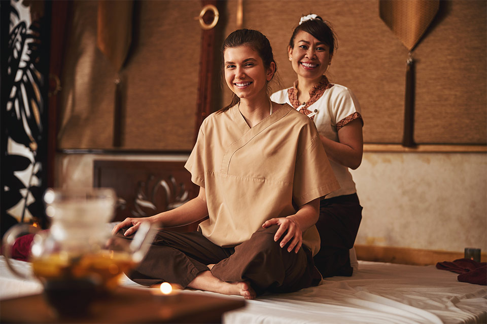 Woman therapist and her client both smile at the camera, with the therapist behind the client sitting cross-legged on a white mattress.