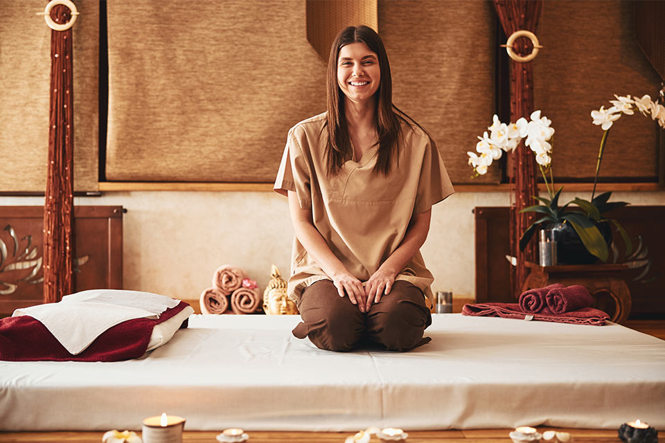 A long-haired smiling woman sits on her knees on a white mattress, facing the camera. Comfortably dressed for Thai massage.