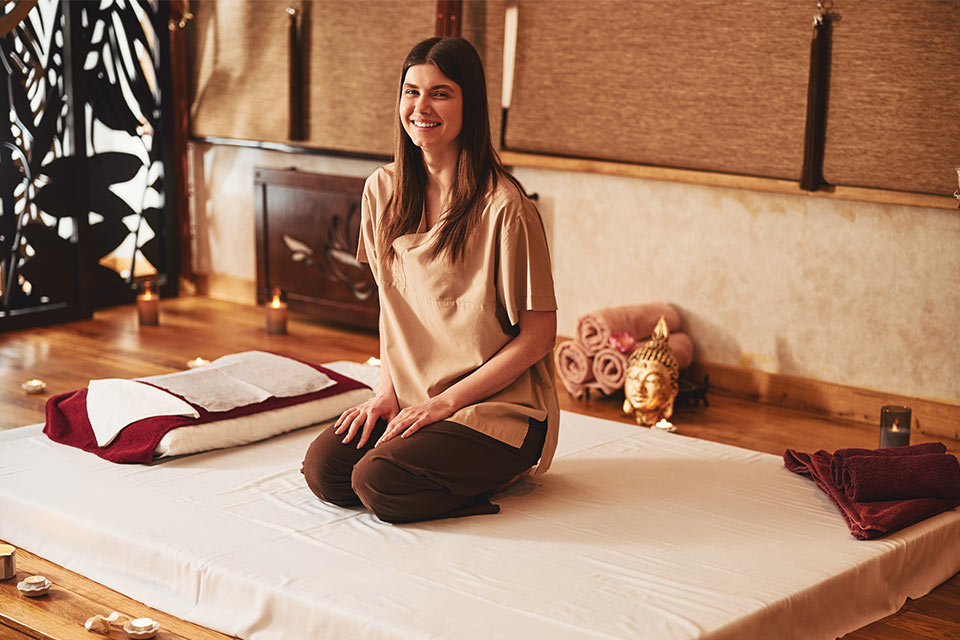 Smiling woman sits on her knees on a white mattress, facing the camera in a three-quarters view, comfortably dressed for Thai massage.